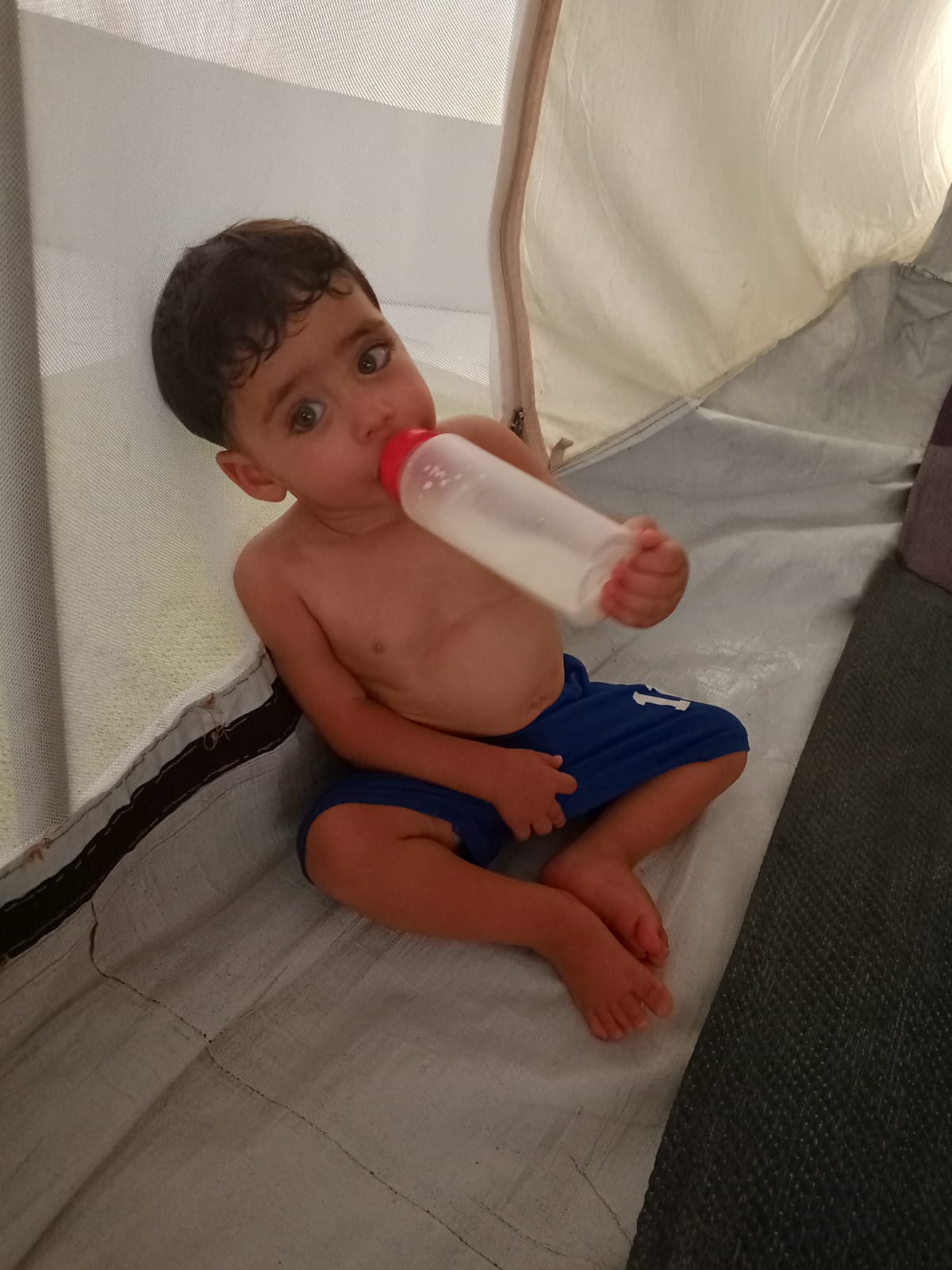 Child sitting inside a tent, holding a bottle